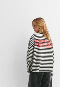 Jonge vrouw met blond haar die een zwart-wit gestreept shirt met lange mouwen draagt met rode tekst op de achterkant en donkere spijkerbroek, zijwaarts kijkend.