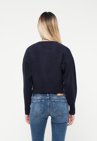 Pulover scurt de culoare bleumarin, cu tricot texturat și mânici largi, asortat cu blugi albaștri care au un design clasic cu cinci buzunare și detalii cusute.