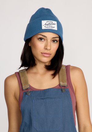 Blue knit beanie with ribbed texture and patch logo, worn with a sleeveless top and denim overalls featuring metal hardware and pocket.
