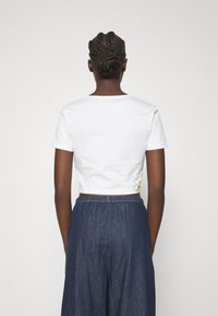 Vit kortärmad t-shirt med avskuren midja som har en liten färgglad blomsterbroderi på baksidan, kombinerad med mörk denimbyxor med vida ben.