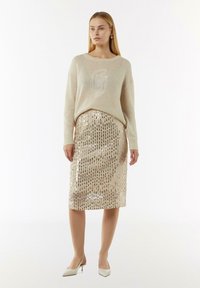 Cremfarbener Pullover mit großem Logo, kombiniert mit einem goldenen Pailletten-Bleistiftrock mit vertikalen Streifen und beigen spitzen Pumps.