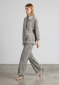 Mujer caminando con un traje de brocado floral gris plata, con una blazer de gran tamaño y pantalones de pierna ancha, combinados con sandalias metálicas de tacón alto.