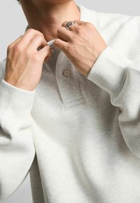Sweatshirt din bumbac de culoare gri deschis, cu guler, placket cu butoane și manșete ribbate. Mâini care ajustează gulerul, punând în evidență un inel de argint pe o mână.