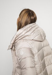 Lichtbeige pufferjack met een gewatteerd ontwerp, grote capuchon en logo op de mouw. Soepele textuur en zichtbare ritssluiting.