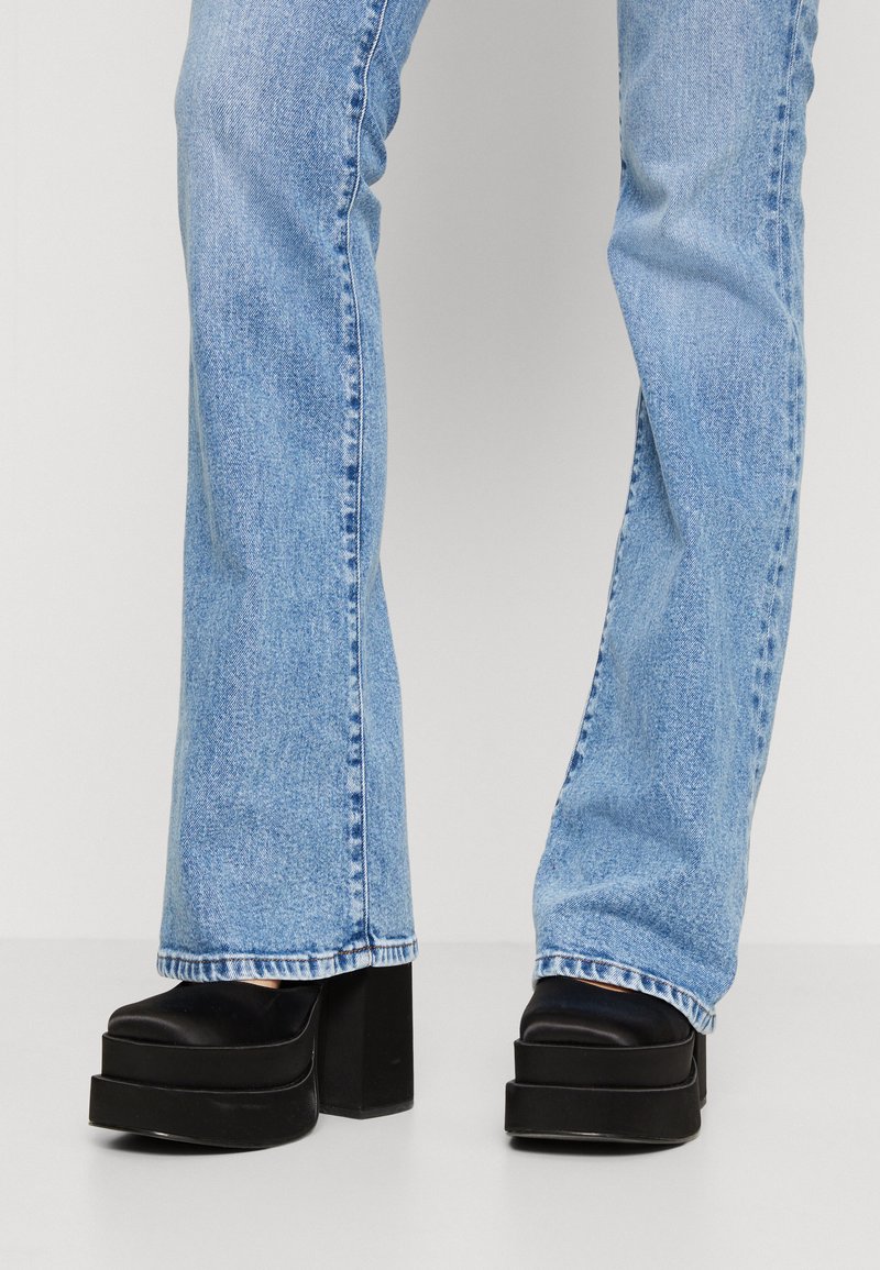 Schwarze Plateaustiefel mit einem breiten, klobigen Absatz und gestuftem Design, kombiniert mit hellblauen Schlagjeans. Der Denim hat eine verduzte Waschung und sichtbare Nähte.