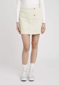 Jupe mini en tweed jaune et blanc avec deux boutons dorés, silhouette droite et tissu texturé. Associée à des baskets blanches et des chaussettes.