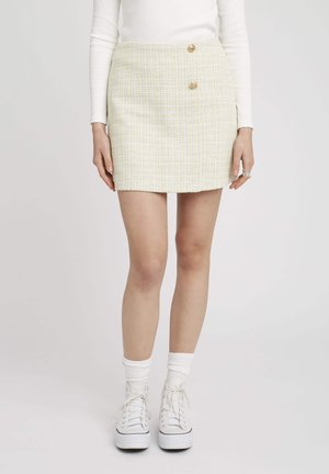 Jupe mini en tweed jaune et blanc avec deux boutons dorés, silhouette droite et tissu texturé. Associée à des baskets blanches et des chaussettes.