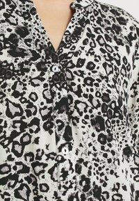 Chemise à imprimé léopard avec une texture soyeuse, comportant un devant à boutons, un col et des motifs noir et blanc sur l'ensemble.