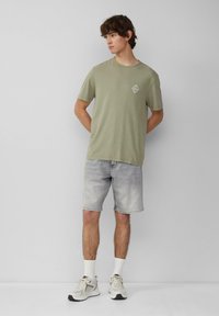 Jasnooliwkowa koszulka z krótkim rękawem z małym nadrukiem, zestawiona z wyblakłymi szarymi szortami dżinsowymi. Białe skarpetki i trampki dopełniają stylizację.