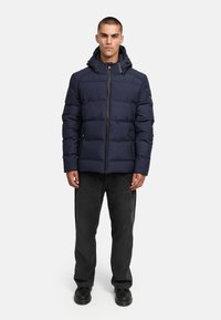 Mörkblå pufferjacka med huva, horisontellt quiltad design, framfickslås med dragkedja, två sidofickor, bärs med svarta manchesterbyxor och kängor.