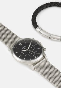 Montre avec bracelet en maille argentée et cadran noir, comportant des index argentés, trois sous-cadrans, et un bracelet en acier inoxydable. Inclut un bracelet en cuir tressé noir.