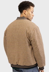 Veste bomber à motif pied-de-poule en marron et beige avec col et poignets côtelés, présentant une coupe décontractée et un tissu texturé.