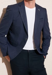 Marinblå blazer i texturerad tyg, med enfaldig knäppning och näsduk i fickan. Bärs över en vit t-shirt med rund hals och mörka byxor.