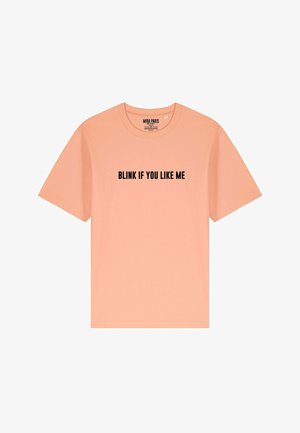 T-shirt en coton de couleur pêche avec des manches courtes et un col rond. L'avant présente un texte noir en gras : "CLIGNE SI TU M'AIMES."