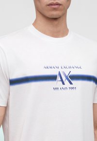 Homme portant un t-shirt blanc avec une bande bleue et le texte « ARMANI EXCHANGE AX MILANO 1991 » sur la poitrine, avec un fond uni.