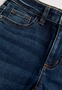 Jean en denim bleu foncé avec surpiqûres orange, montrant la poche avant, la petite poche à monnaie, la passants de ceinture et la ceinture.