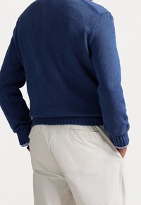 Pull en maille bleu marine avec un ourlet et des poignets côtelés, présentant une finition texturée. Associé à un pantalon beige clair avec des poches arrière.