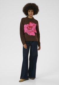 Brown knit sweater featuring a large pink floral design on the front, paired with dark blue flared jeans and beige footwear.