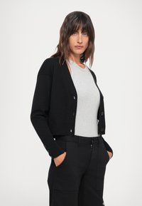 Svart kort cardigan med rund hals, knappelukking og ribbede mansjetter, båret over en lys grå tettsittende topp og sorte bukser.