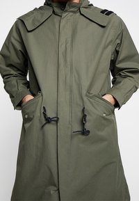 Cappotto lungo verde khaki realizzato in tessuto leggero, con colletto alto, grandi tasche e cordini regolabili in vita e all'orlo.