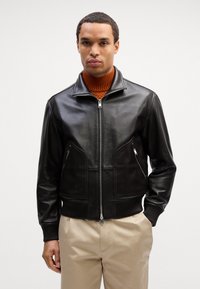 Giubbotto bomber in pelle nera con zip frontale, due tasche frontali angolari, polsini e orlo a coste, indossato sopra un maglione a collo alto arancione.