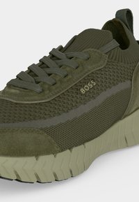 Baskets vert olive en tissu tricoté texturé, détails en daim, semelle épaisse rainurée et logo "BOSS" sur le côté près des lacets.