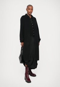 Cappotto lungo nero con colletto e bottoni, abbinato a una maglia nera e una gonna voluminosa. Stivali lucidi bordeaux e una borsa nera.