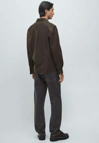 Mężczyzna stojący bokiem, ubrany w ciemnobrązową koszulę, czarne dżinsy i czarne buty, na tle jednolitego jasnoszarego tła.