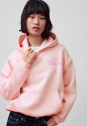 Mladá žena s krátkymi tmavými vlasmi, oblečená v svetloružovej mikine "Barbie Fairytopia" a tmavých rifle, stojaca pred sivým pozadím.