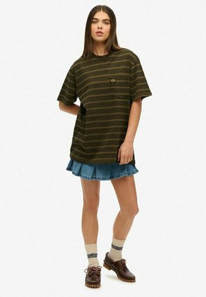 T-shirt surdimensionné vert et brun à rayures avec une poche sur la poitrine, associé à une jupe en denim. Le mannequin porte des chaussures brunes et des chaussettes rayées.