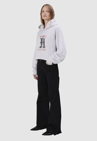 Kobieta stoi, ubrana w jasnoszary krótki kaptur z nadrukiem butów oraz czarne, szerokie spodnie, na jasnym, jednolitym tle.