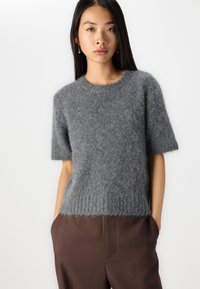 Maglione grigio a maniche corte in materiale soffice con scollatura rotonda e orlo a costine, abbinato a pantaloni marroni. Aspetto testurizzato e morbido.