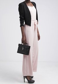 Mustat lyhennetyt bleiserit vaaleanpunaisen haalarin päällä, jossa on sidottu vyötärö. Kädessä musta käsilaukku metallikannella. Mustat korkokengät.