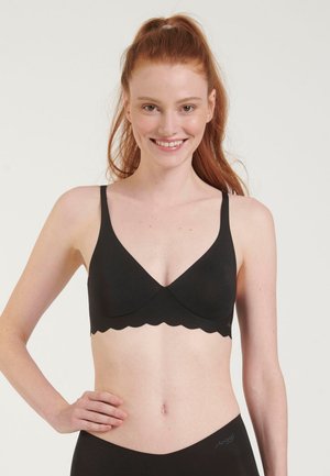 Uśmiechnięta kobieta z rudymi włosami w wysokim kucyku, ubrana w czarny stanik z falistym brzegiem i pasujące czarne majtki na białym tle.