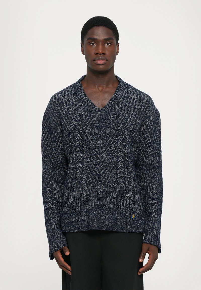 Junger schwarzer Mann, der nach vorne schaut, trägt einen dunkelblauen strukturierten V-Ausschnitt-Pullover und schwarze Hosen vor einem schlichten weißen Hintergrund.