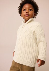 Pull en tricot couleur crème avec un col montant, présentant des motifs en côtes et des poignets côtelés, associé à un pantalon beige.