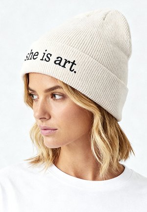 Cream-colored ribbed beanie with the text "she is art." in black. Features a folded cuff and soft texture. Worn with a white shirt.