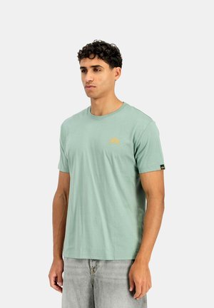 Mann mit kurzen lockigen Haaren trägt ein hellgrünes kurzärmeliges T-Shirt und hellgraue Jeans, steht vor einem schlichten weißen Hintergrund.