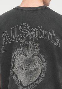 Μαύρο βαμβακερό μπλουζάκι με γραφικό σχέδιο που περιλαμβάνει μια καρδιά, διακοσμητικά στοιχεία που μοιάζουν με λουλούδια και το κείμενο "All Saints" σε ασημί γράμματα.