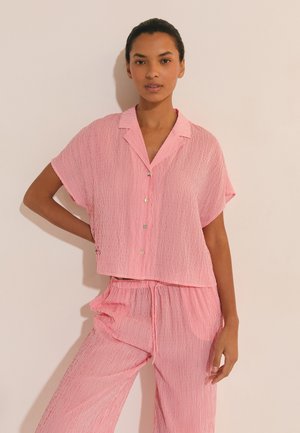Femme portant une chemise boutonnée à manches courtes ample à rayures roses et blanches et un pantalon ample assorti, sur un fond uni.