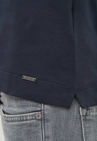 T-shirt en coton bleu marine avec une �étiquette en métal "JOOP!" au bas de l'ourlet, présentant une fente sur le côté et une texture lisse. Un denim gris est visible.