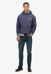 Man in een blauwe "dry SUPER" hoodie, een geruite shirt, jeans, zwarte laarzen en een horloge, staat tegen een witte achtergrond.