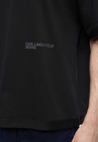 Μαύρο μπλουζάκι με το κείμενο "KARL LAGERFELD JEANS" τυπωμένο με μικρά γκρι γράμματα στο στήθος, φορεμένο από άτομο με ορατό χέρι και σκούρο τζιν.