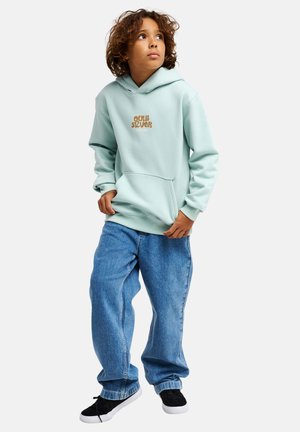 Sudadera con capucha verde claro con bolsillo frontal y texto de logo marrón, combinada con vaqueros azules holgados y zapatos negros. Estilo informal de ropa urbana.