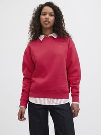 Sweatshirt rouge avec des poignets côtelés, porté sur une chemise blanche à rayures avec col. Un pantalon en denim foncé complète le look. Cheveux bouclés visibles.