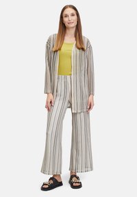 Gestreepte stoffen set met een lange cardigan met wijde mouwen, gecombineerd met een loose-fit broek. Kleuren zijn beige, grijs en olijfgroen. Zwarte sandalen.