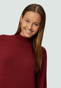 Burgunderfarbener Strickpullover mit hohem Kragen, langen Ärmeln und weicher Textur, in einer Nahaufnahme auf neutralem Hintergrund präsentiert.
