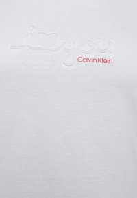 Valkoinen puuvillapaita, jossa on kohotettu teksti "i❤️you" ja "Calvin Klein" vaaleanpunaisella kirjaimella, tasainen pinta ja yksinkertainen muotoilu.