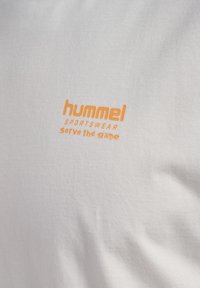Orange "hummel SPORTSWEAR serve the game" logo på hvidt stof.