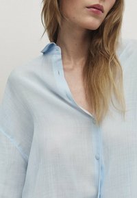 Ljusblå knapp-up skjorta gjord av ett genomskinligt, lätt tyg; har krage, lösa ärmar och subtil textur.